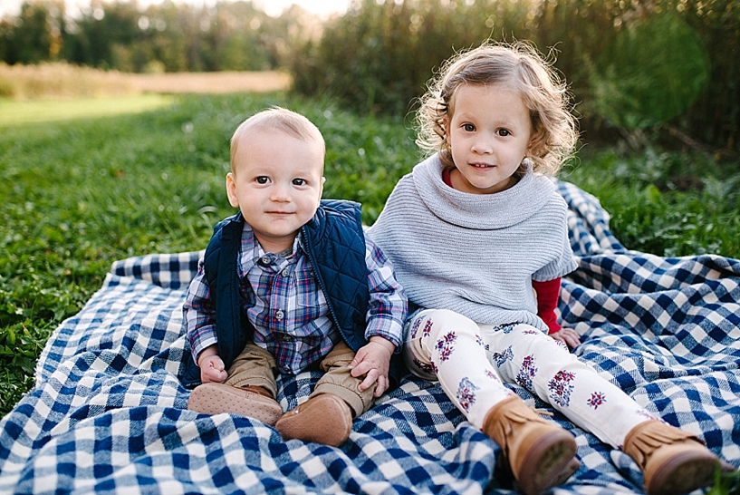 baby boy in blue plaid shirt and blue vest and older sister in grey poncho and floral leggings sitting on blanket by field