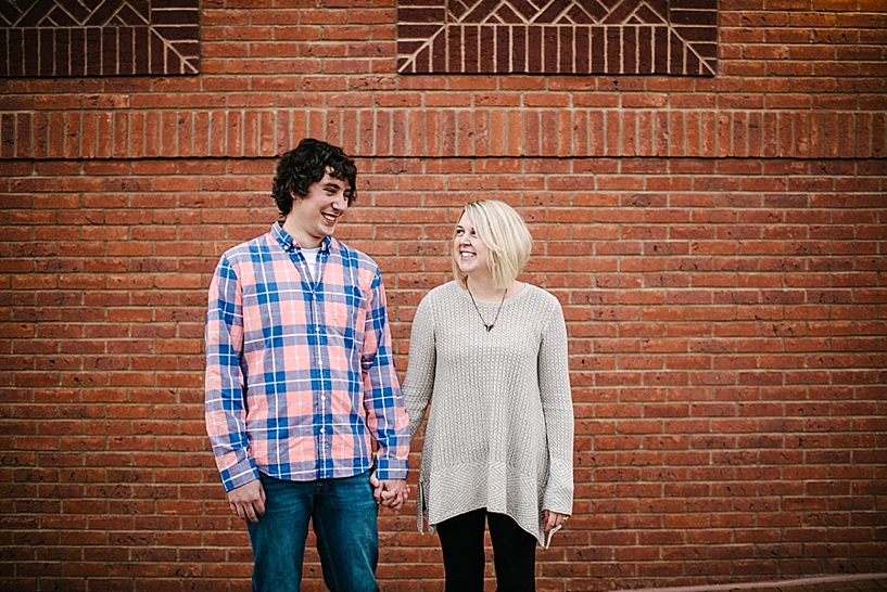 girl in oversized sweater and guy in pink and blue plaid shirt standing in front of brick wall holding hands
