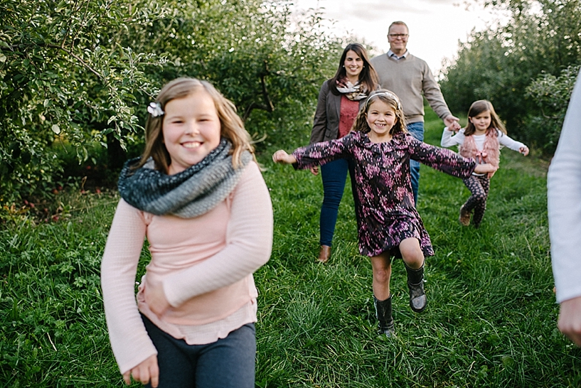 kids running and laughing in apple orchard with parents walking behind them smiling