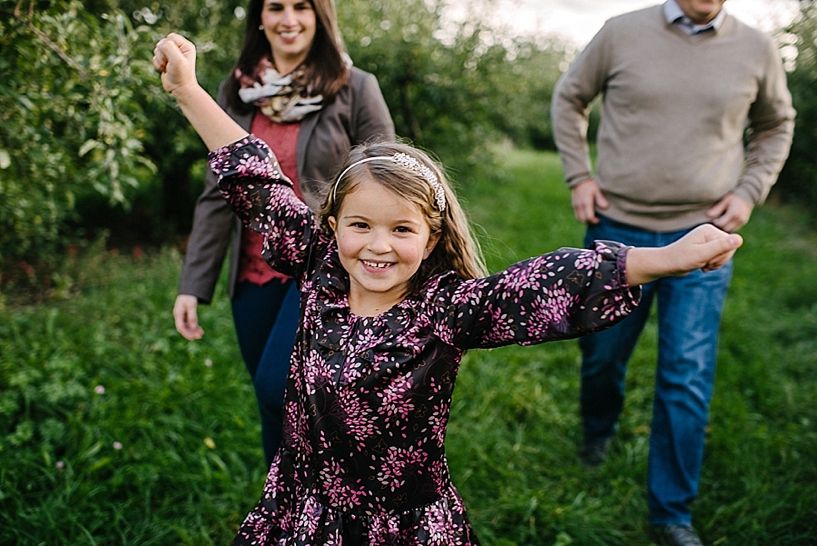 little girl smiling with outstretch arms in apple orchard with parents walking behind her smiling