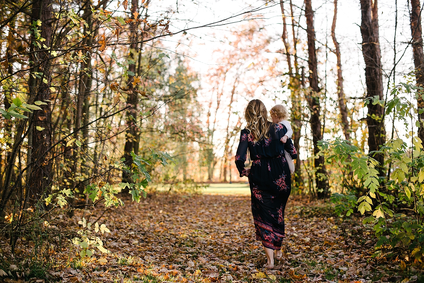 mother in navy floral print dress holding daughter in arms and walking through the woods
