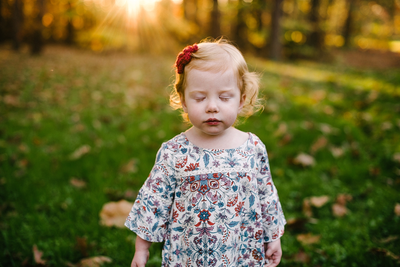 toddler redhead girl wearing floral print shirt standing with eyes closes and sun shining behind her