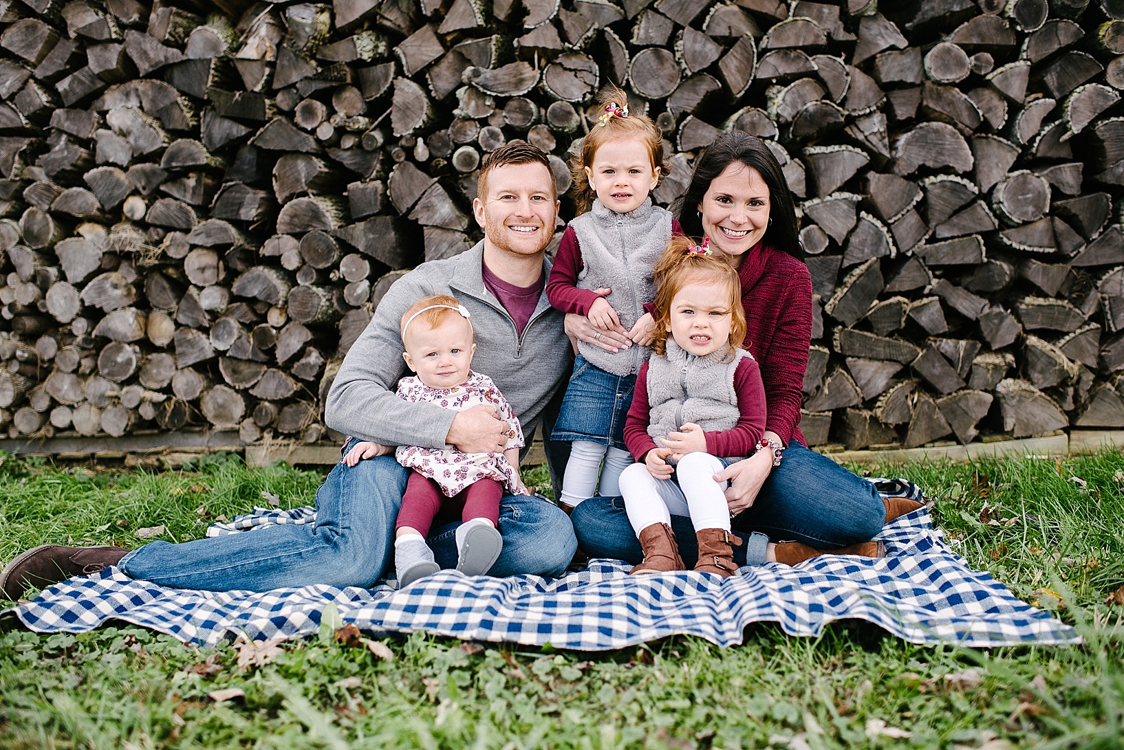 parents with three young daughters sitting on blue plaid blanket in front of wood pile