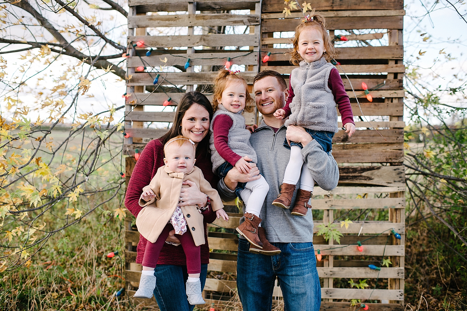 parents holding three daughters dressed in grey and maroon in front of DIY pallet wall