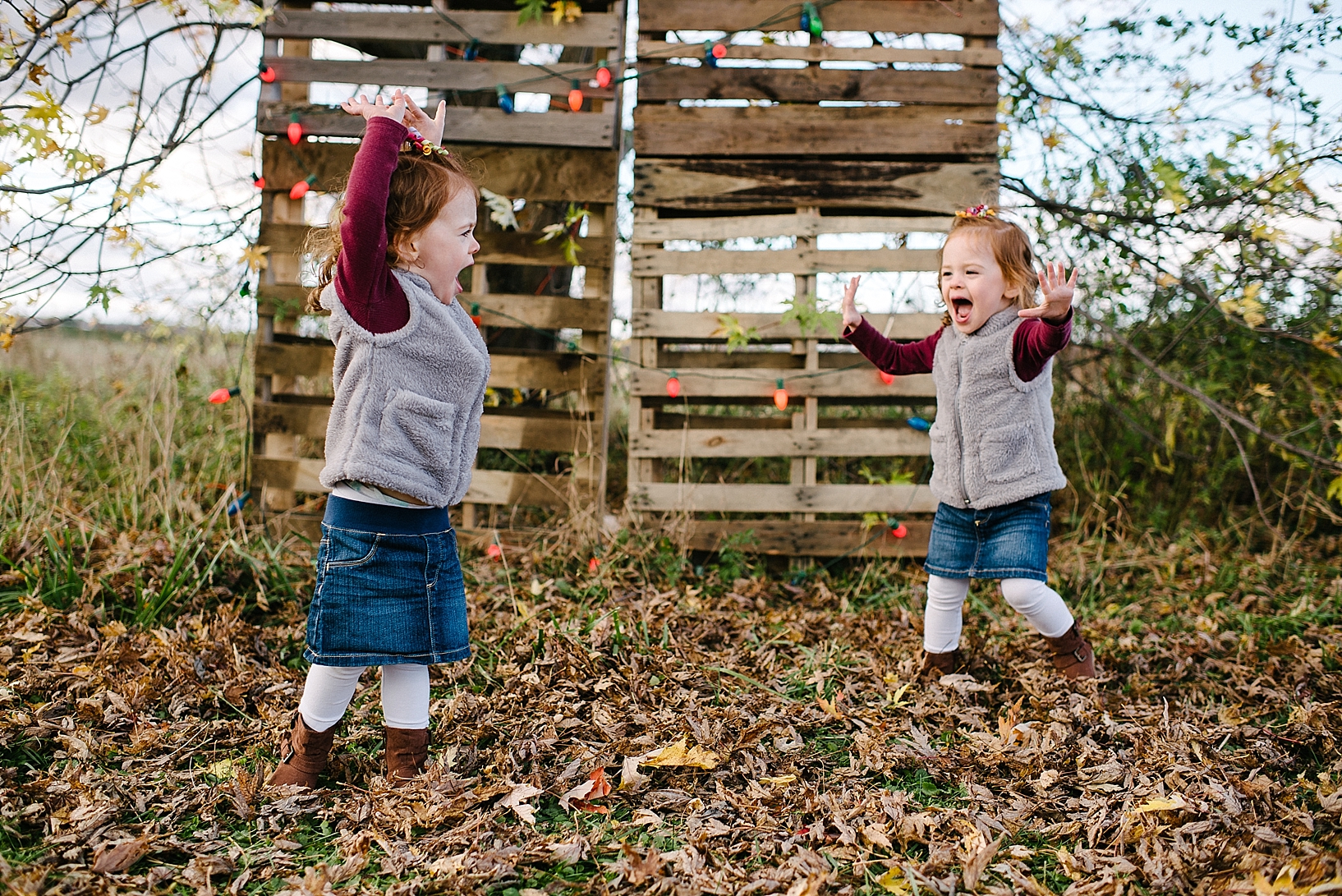 twin toddler redheads wearing grey vests and denim skirts dancing in front of wood pallet wall