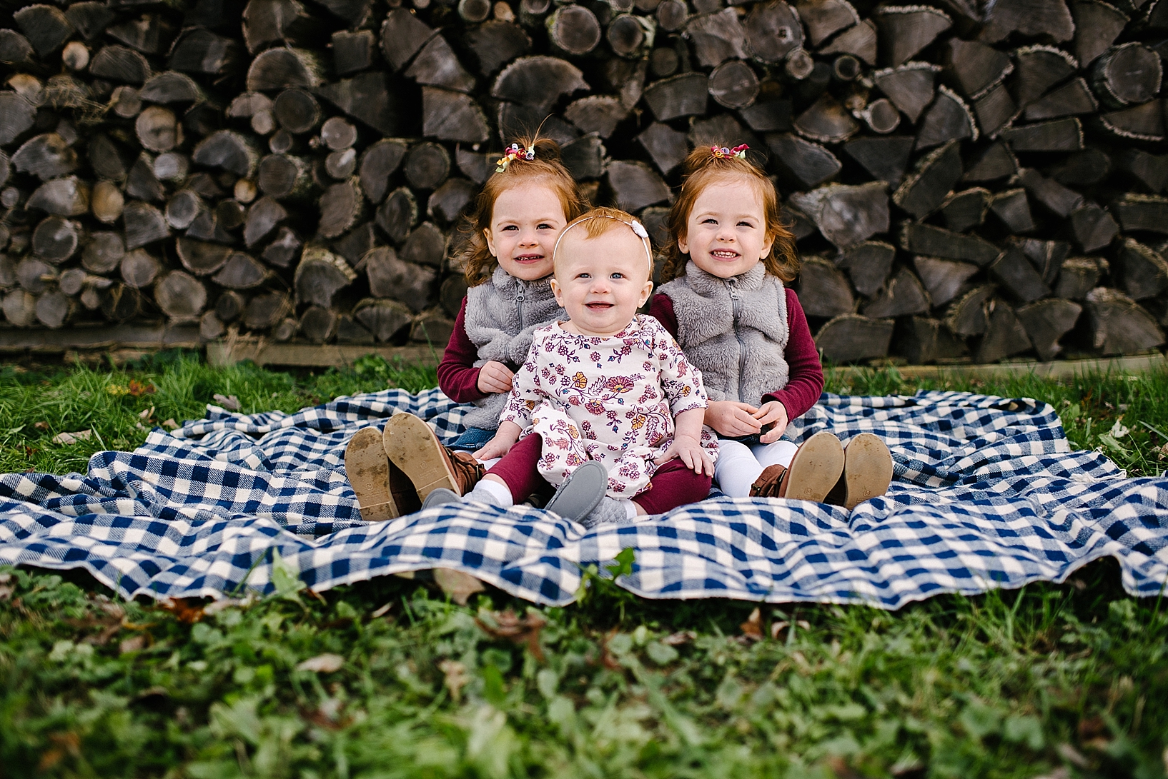 three redhead toddlers sitting on blue plaid blanket in front of wood pile