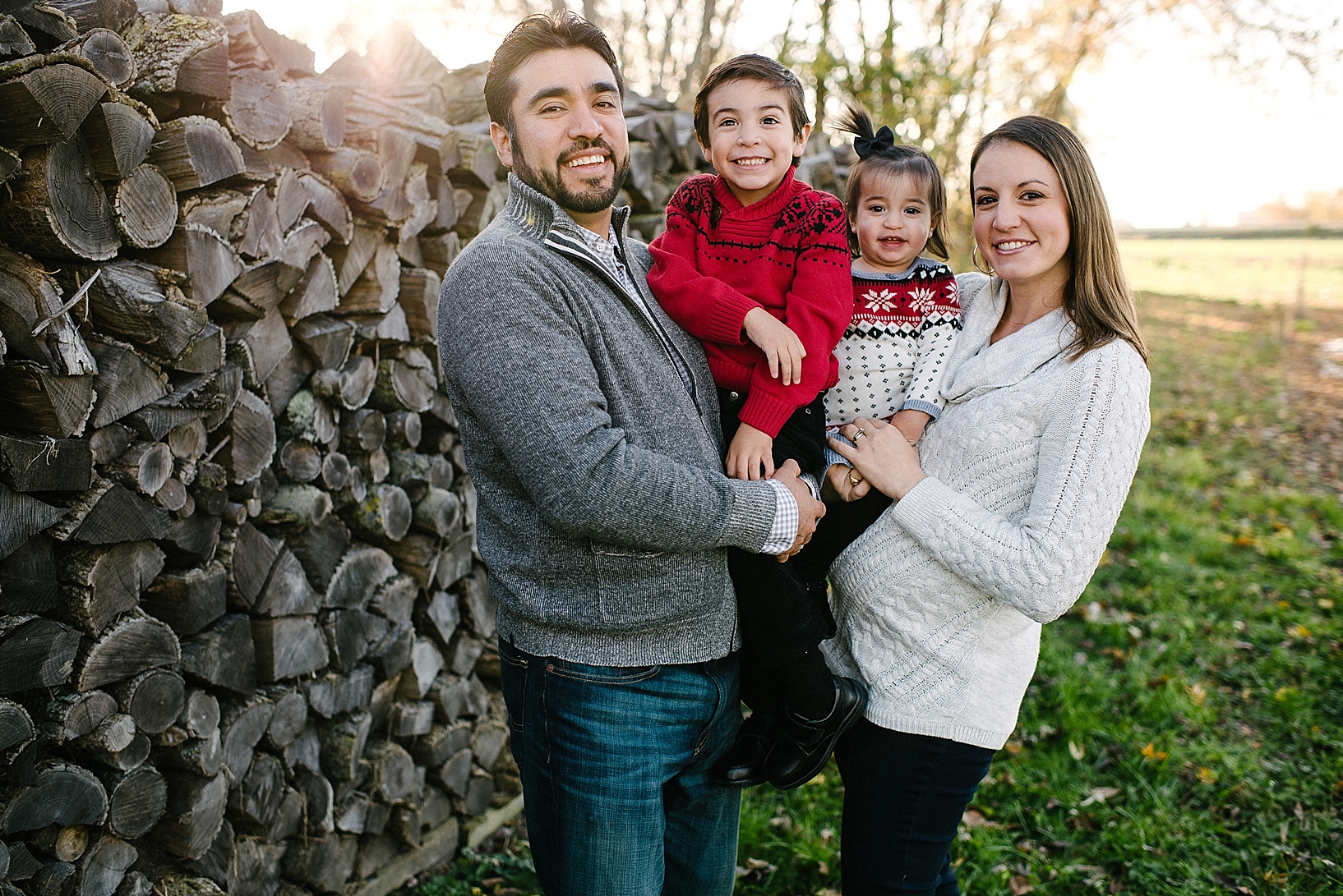 family in grey, cream, and red sweaters standing in front of log pile