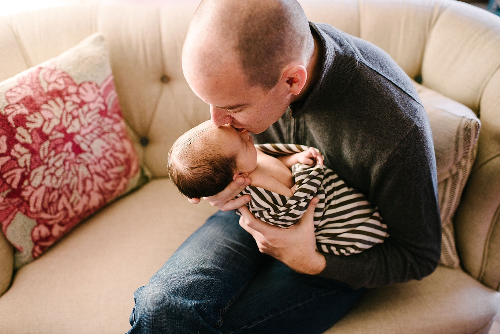 dad in grey sweater kissing newborn son wrapped in brown striped blanket on forehead