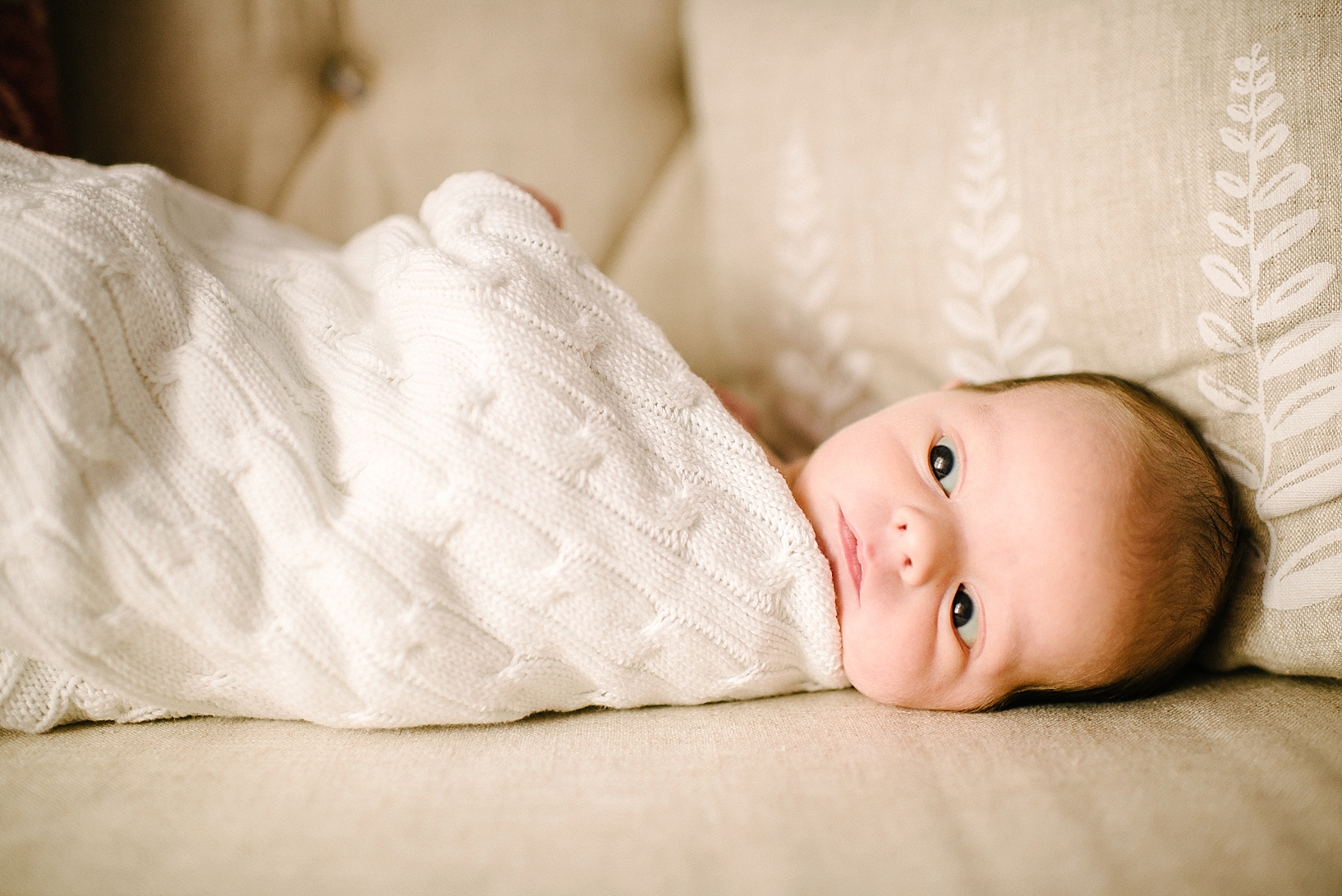 newborn baby wrapped in cable knit blanket on tan sofa