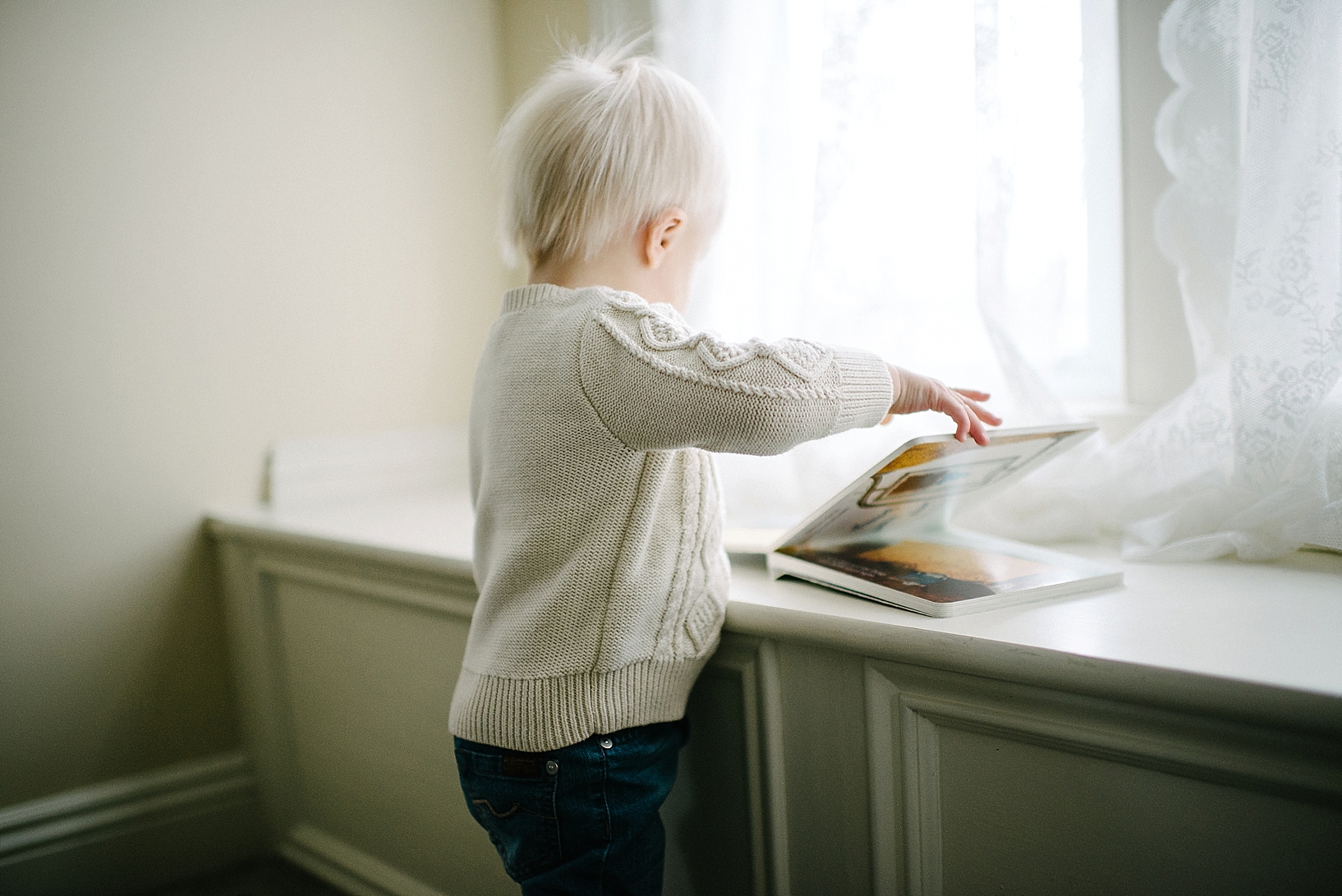 toddler boy with blonde hair and cable knit sweater reading book by window