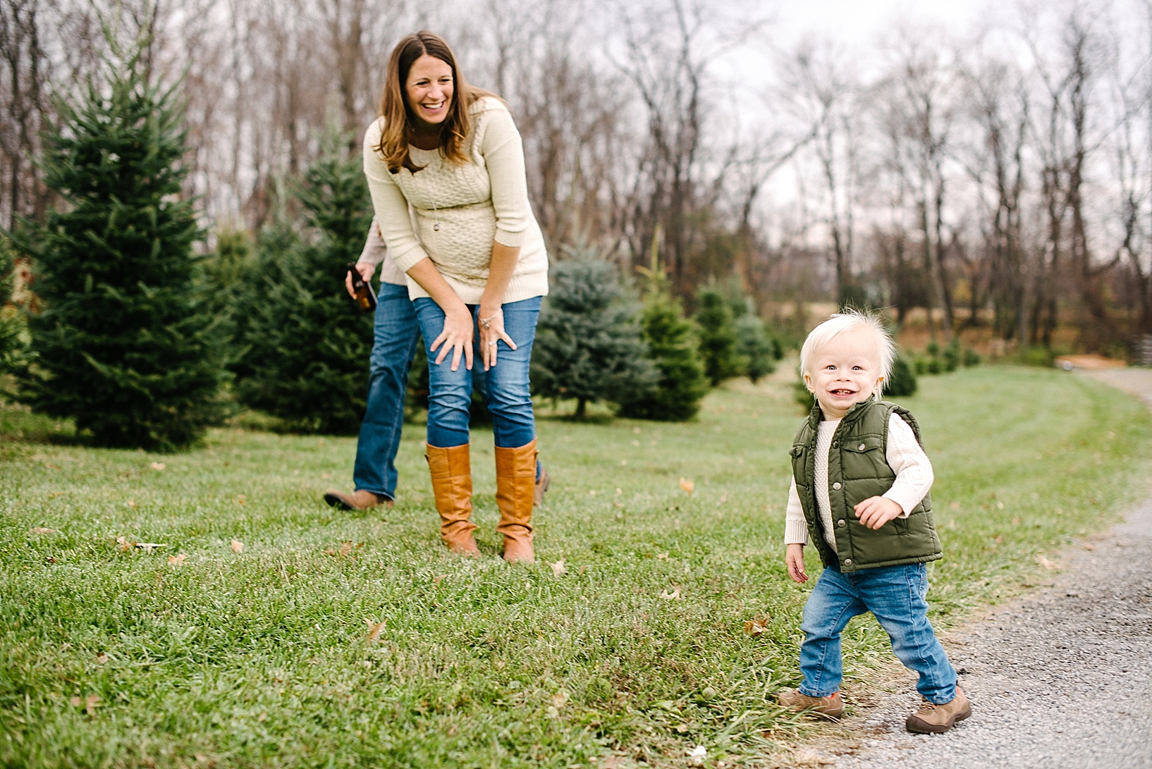 toddler boy in jeans and green vest running with expectant mother in cable knit sweater laughing in the background