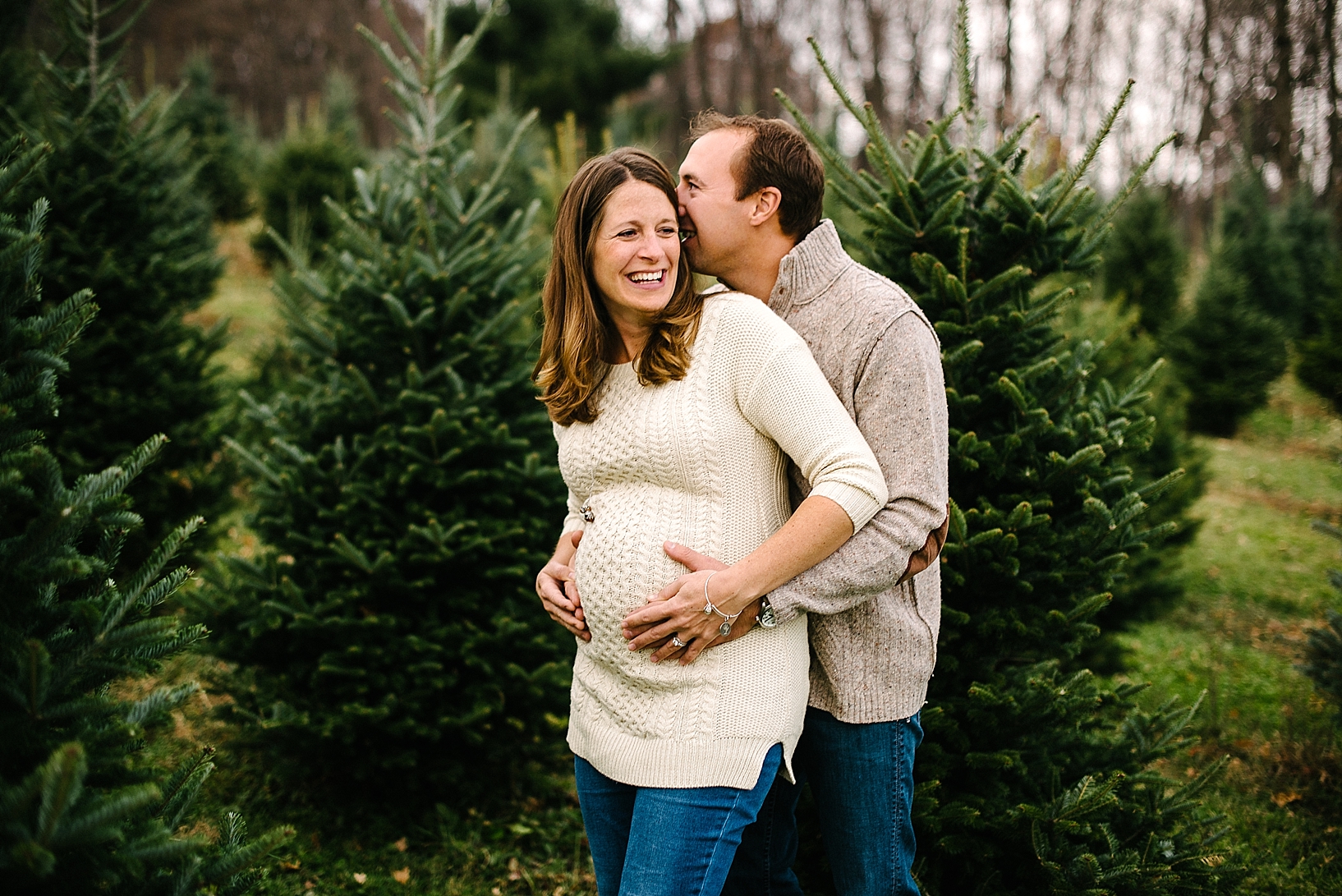 expectant mother wearing cable knit sweater laughing while husband whispers in her ear with arms wrapped around her belly standing among pine trees