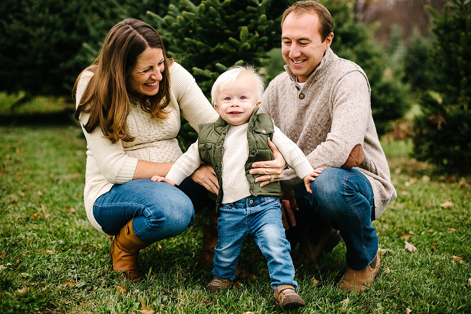 parents in cable knit sweaters kneeling next to toddler son in green vest and jeans in front of pine trees