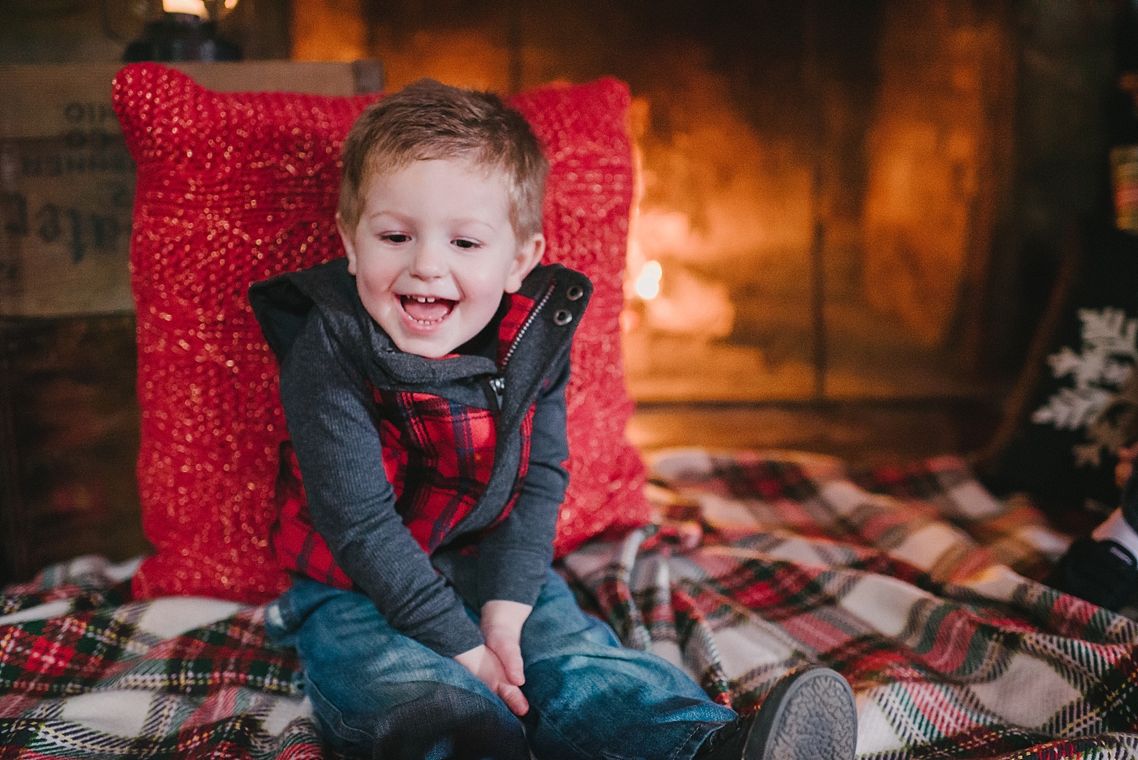 little boy sitting on plaid blanket in front of fireplace laughing with hands in his lap