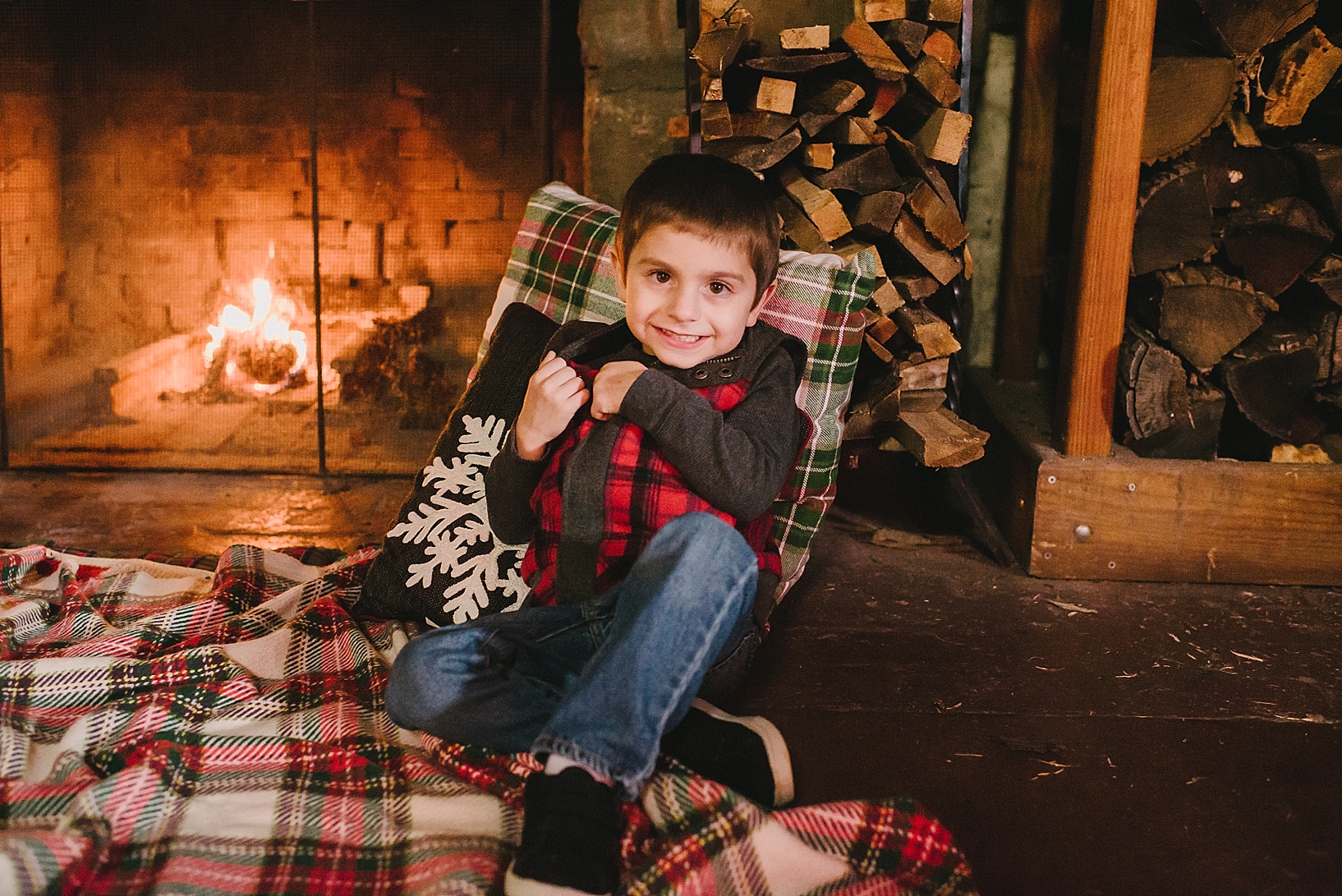 little boy sitting on plaid blanket leaning on pillow by wood pile in front of fireplace