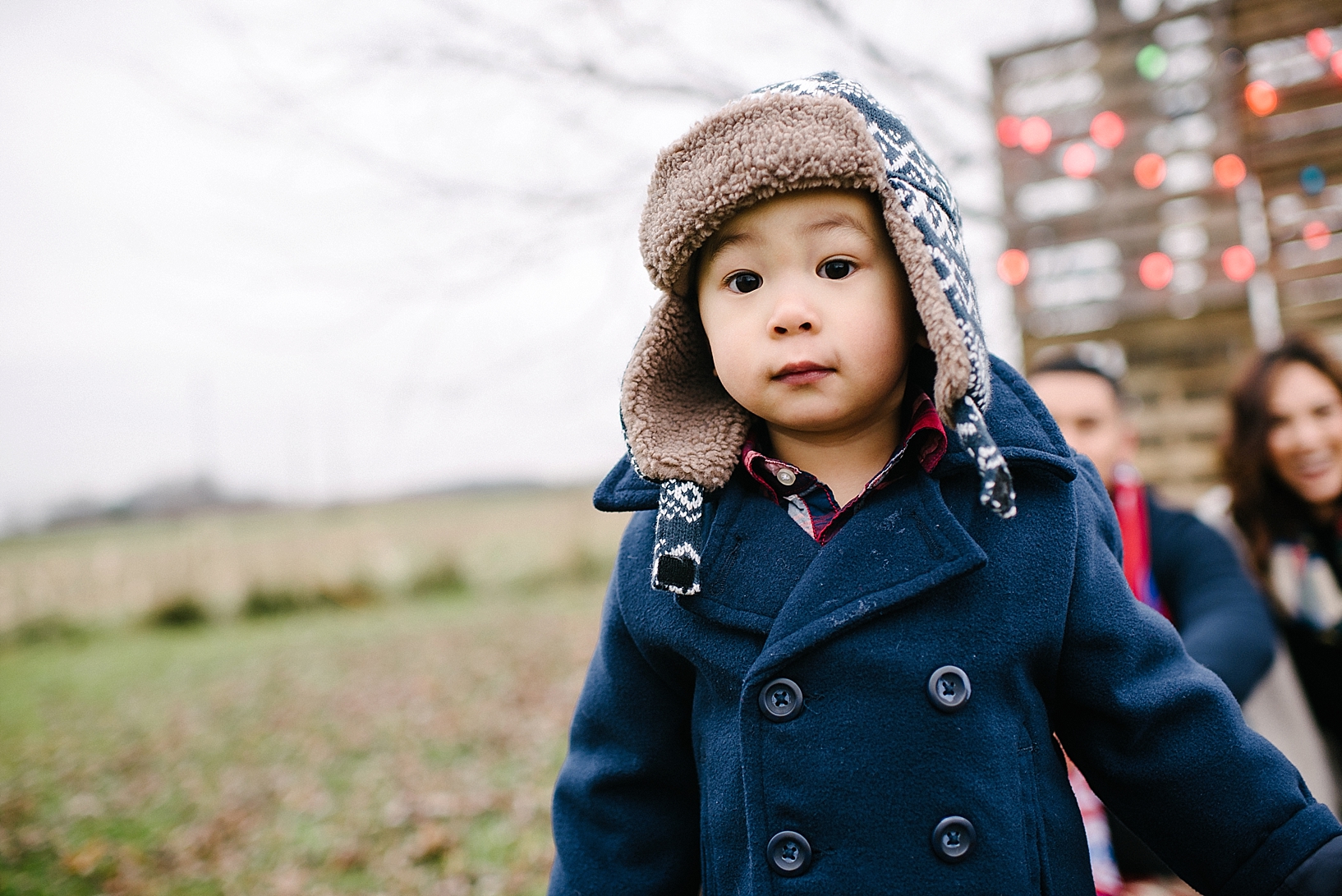 toddler boy wearing navy blue pea coat and warm fuzzy hat with parents smiling in the background