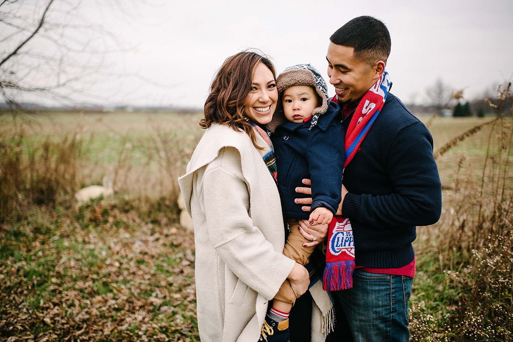 family with toddler son dressed in winter coats standing in country field