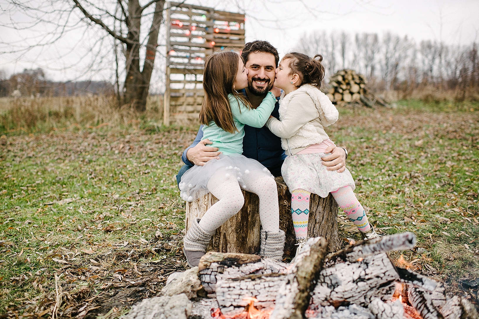 young girls dressed in colorful winter outfits sitting on tree stumps by campfire kissing their dad's cheeks