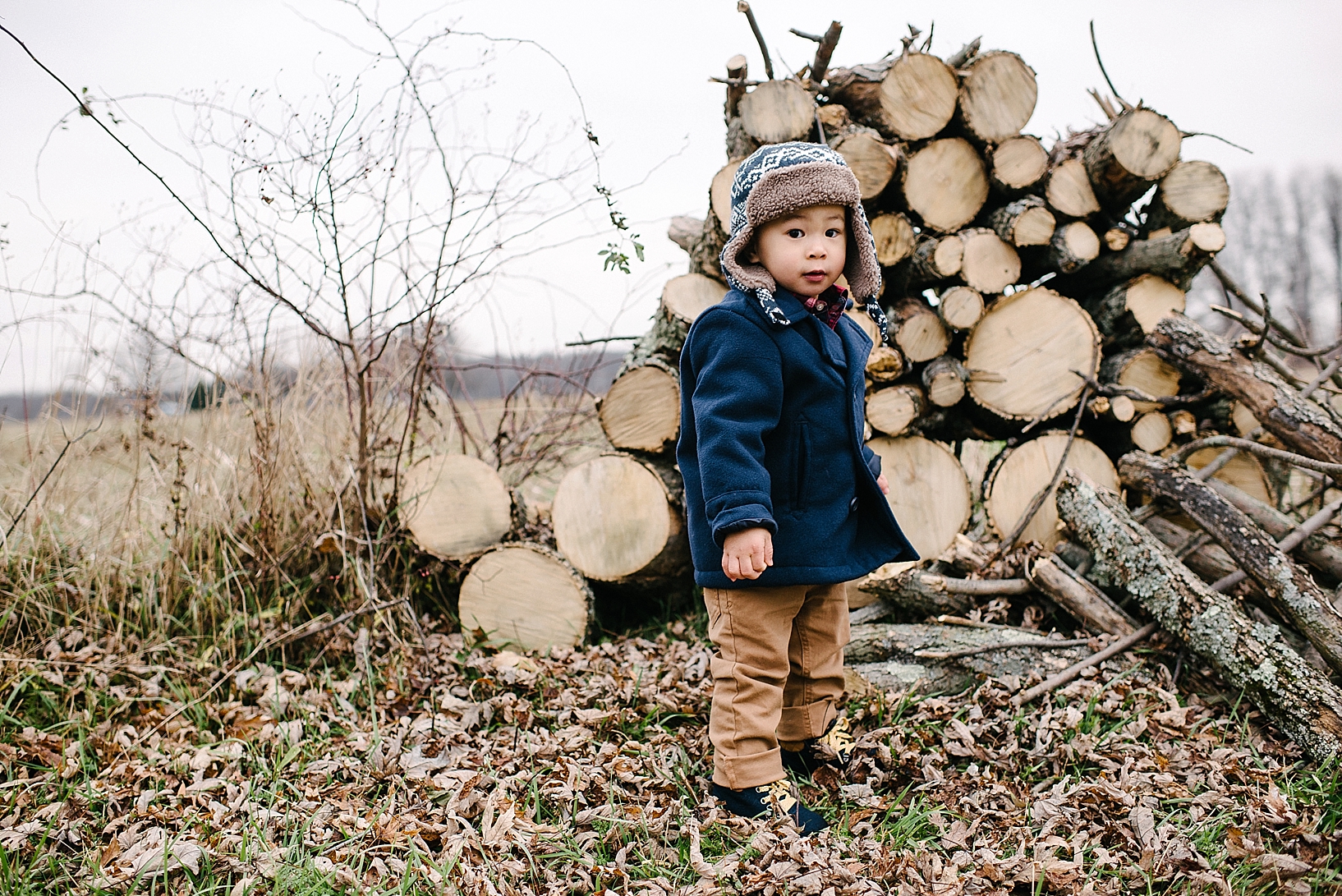 toddler boy wearing navy pea coat and khakis standing in front of log pile