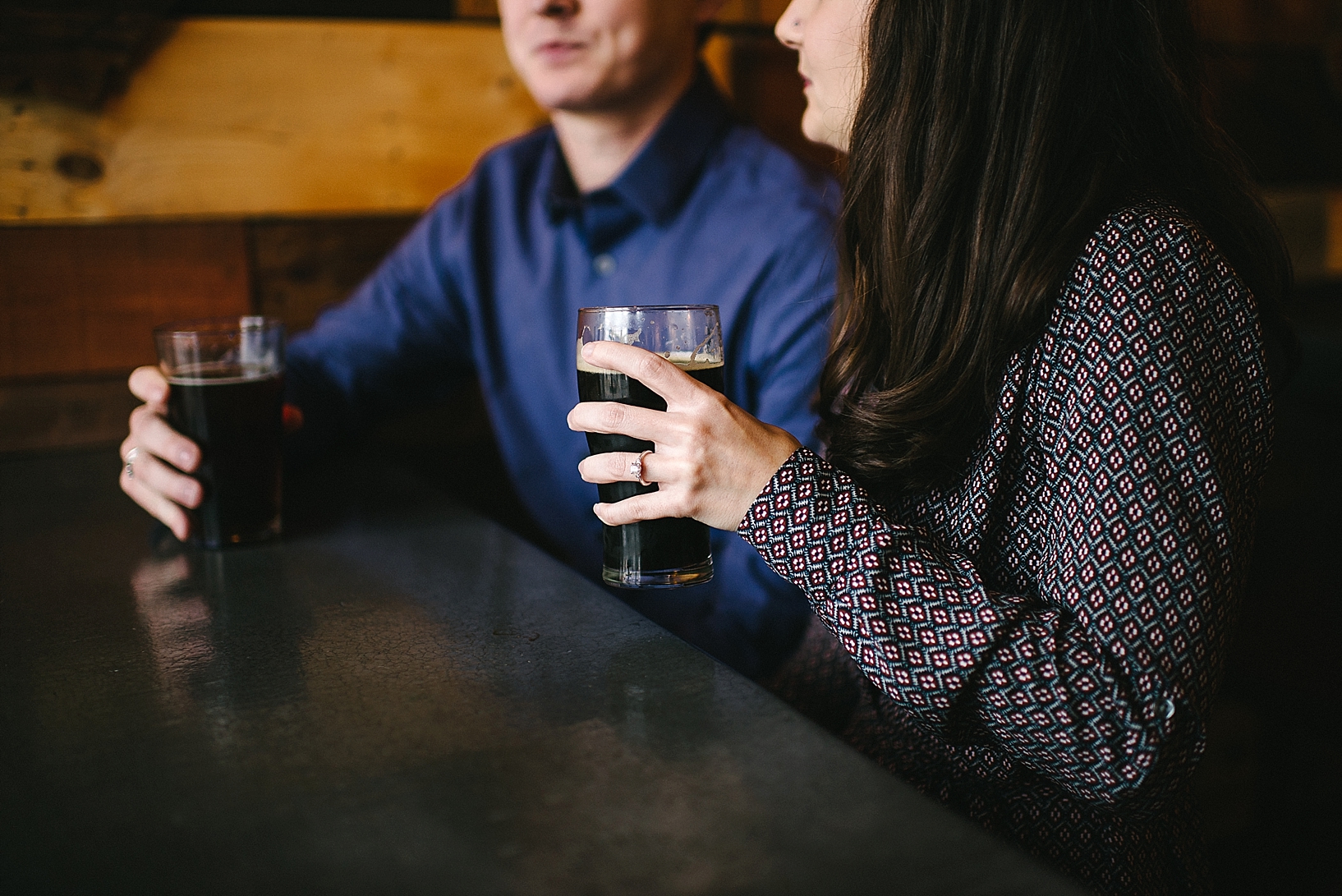 woman's hand wearing diamond engagement ring holding craft beer sitting next to fiance in blue shirt