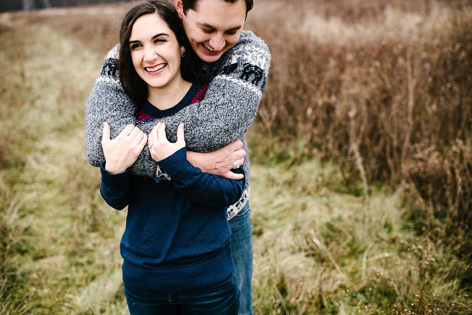 guy in grey sweater standing behind girl in blue sweater hugging and laughing