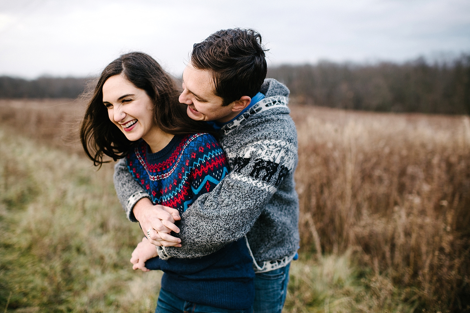 guy in grew sweater hugging woman in blue sweater from behind and laughing