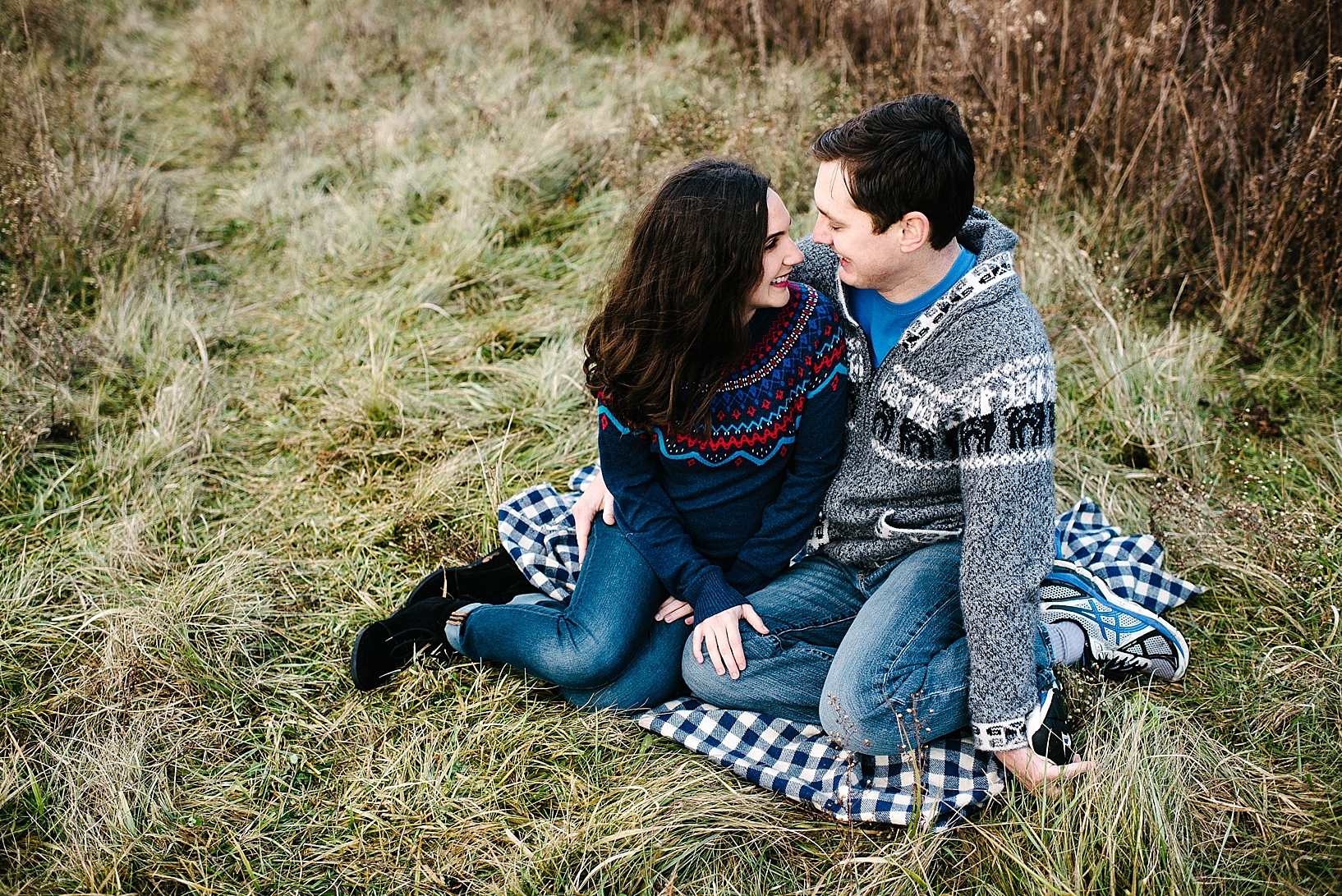 guy in grey sweater and girl in blue sweater sitting on blue checked blanket in the grass