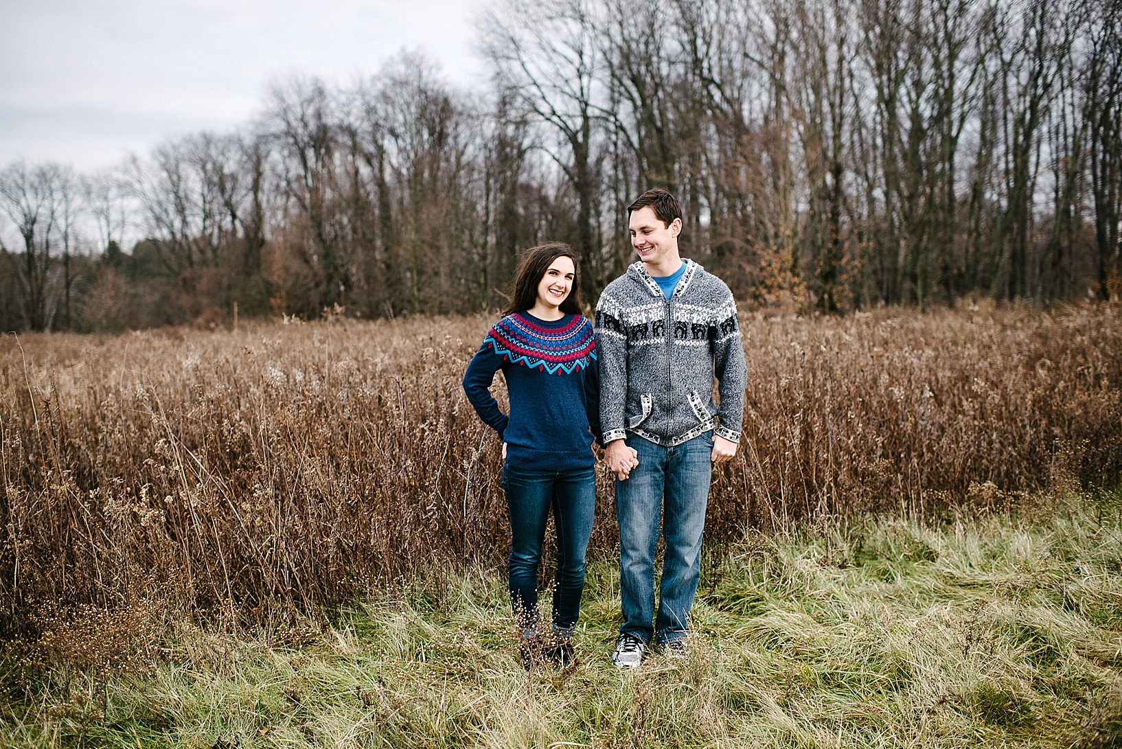 guy in grey sweater and girl in blue sweater standing side by side in field in winter