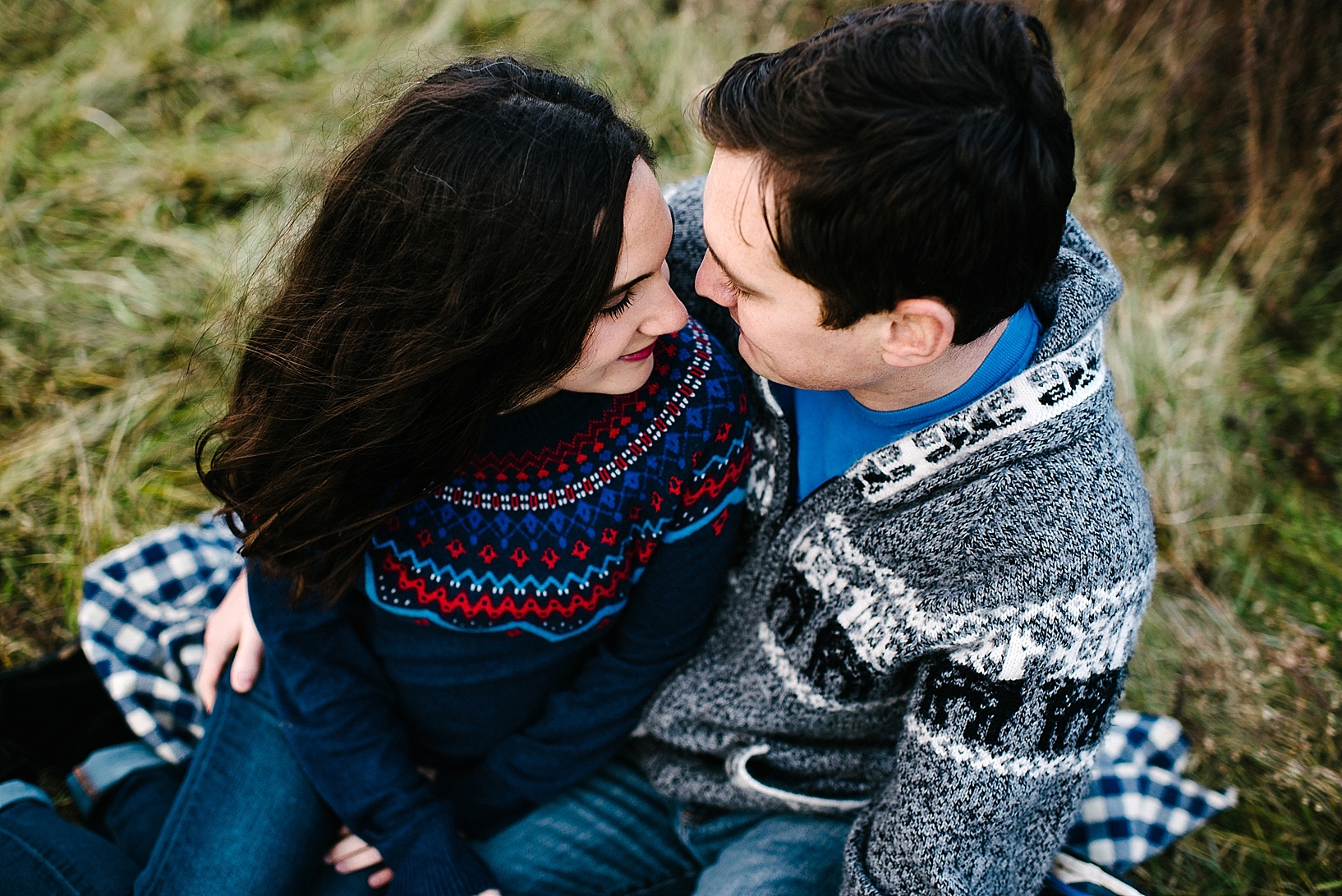 guy in grey sweater and girl in blue sweater sitting on blue checked blanket in middle of field