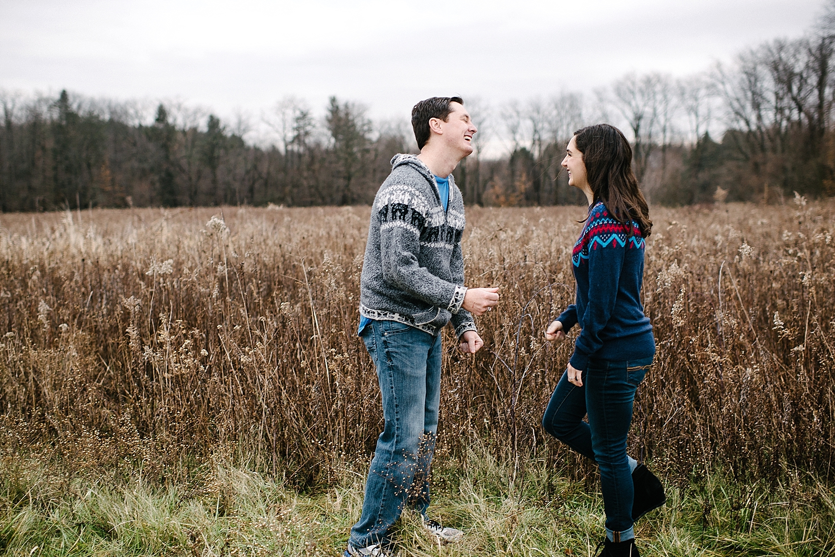 guy in grey sweater and girl in blue sweater standing facing each other in field laughing