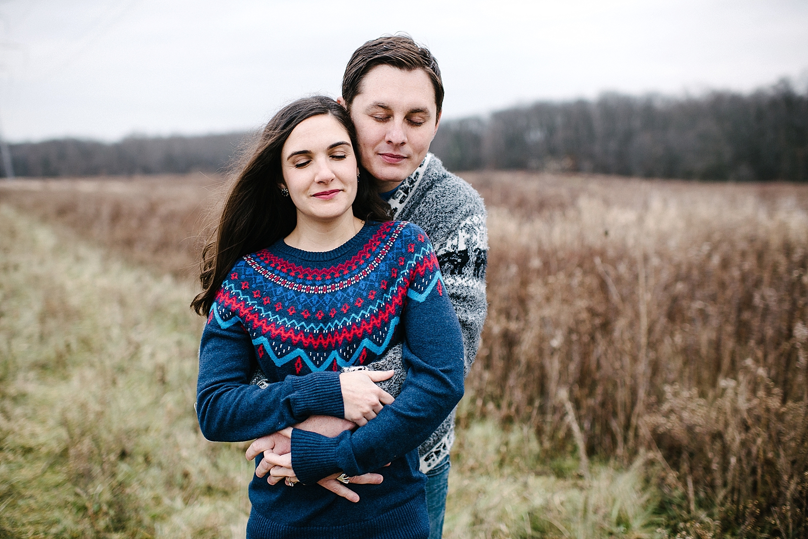 guy in grey sweater and girl in blue sweater standing in field with arms wrapped around each other and eyes closed