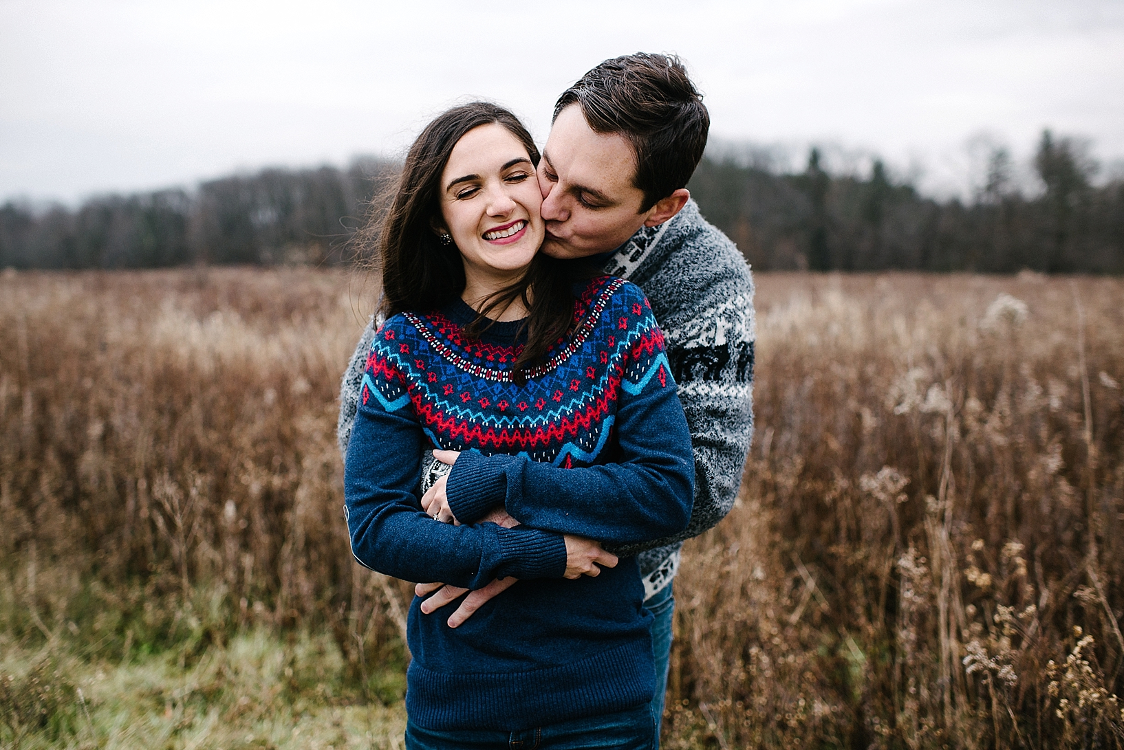 guy in grey sweater kissing girl in blue sweater on the cheek while she smiles