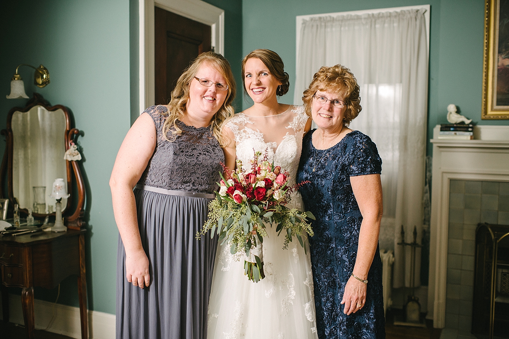 bride holding bouquet standing next to mother in navy dress and sister in gray lace dress