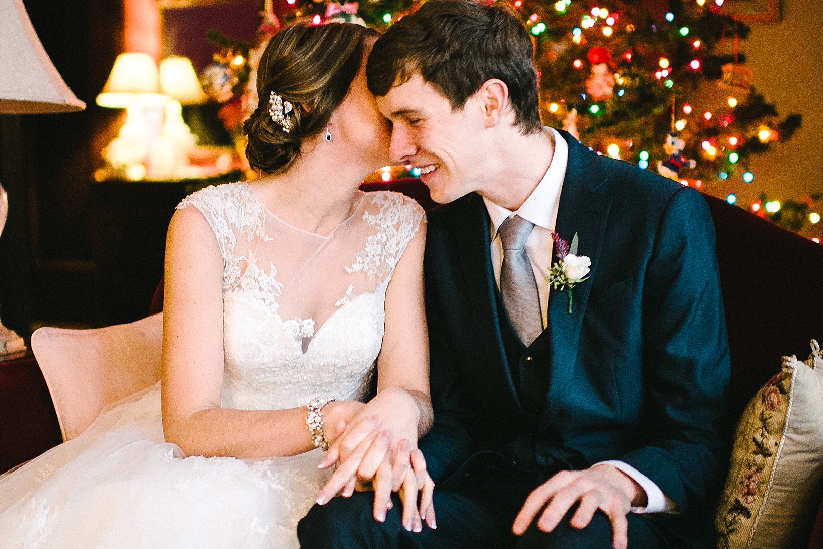 bride and groom sitting on couch in front of Christmas tree whispering in each other's ear