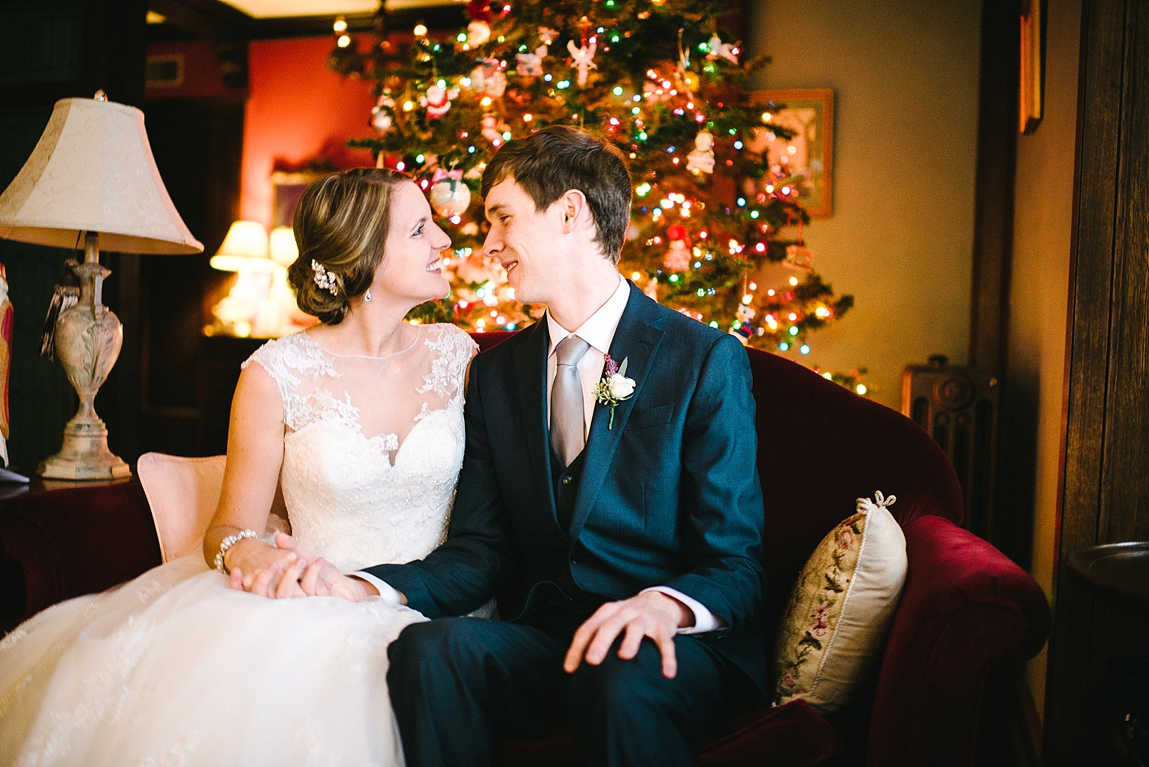 bride and groom sitting on couch in front of Christmas tree smiling at each other