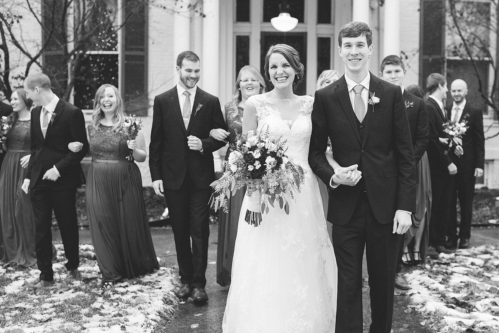 bride and groom walking hand in hand up snowy sidewalk with bridal party walking behind