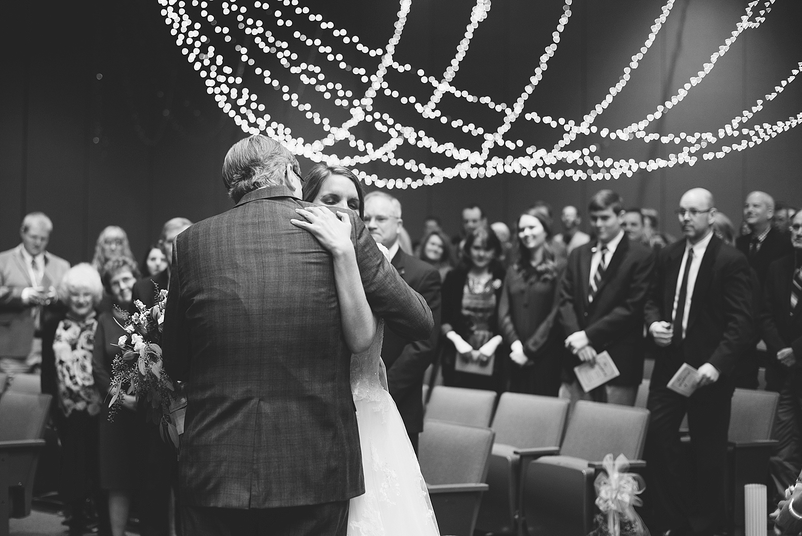 father hugging bride at front of church with string lights in the background