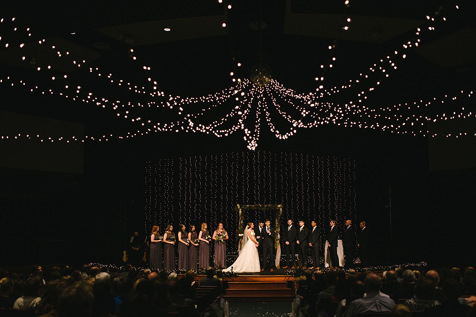 wedding party standing on church stage with white twinkle lights hanging from ceiling