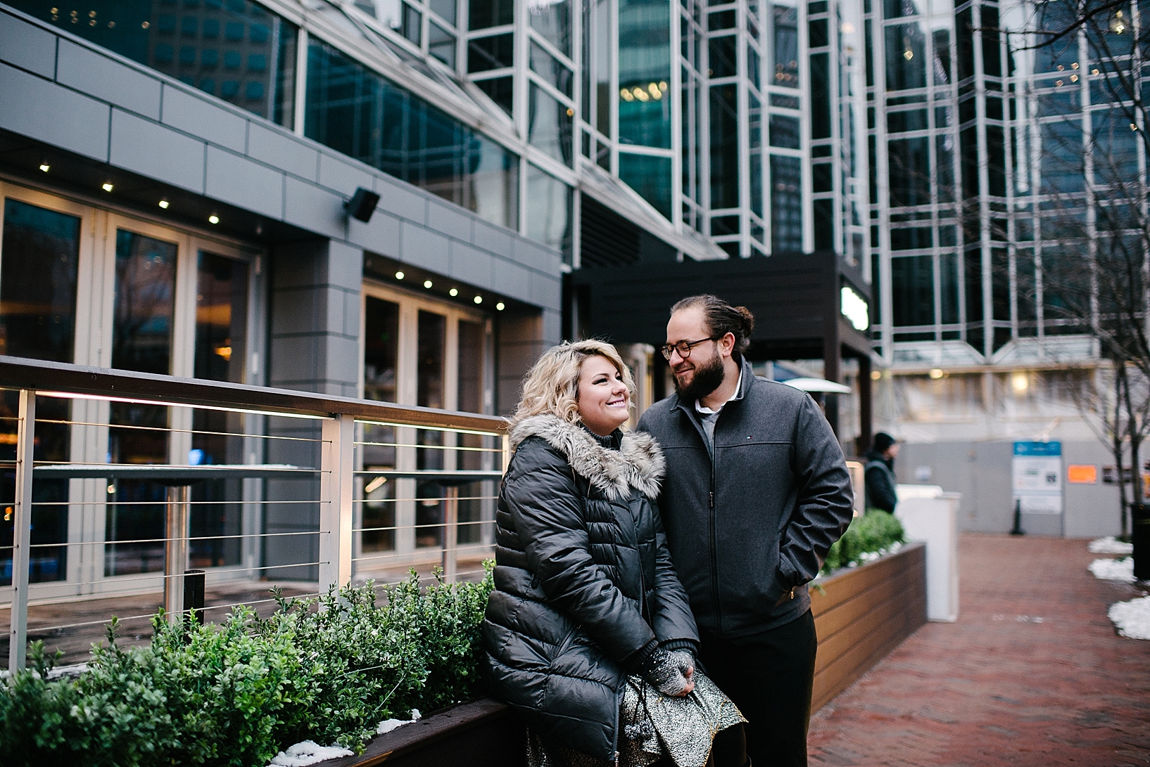 blonde woman wearing puffy winter coat sitting on ledge looking up at fiance with man bun
