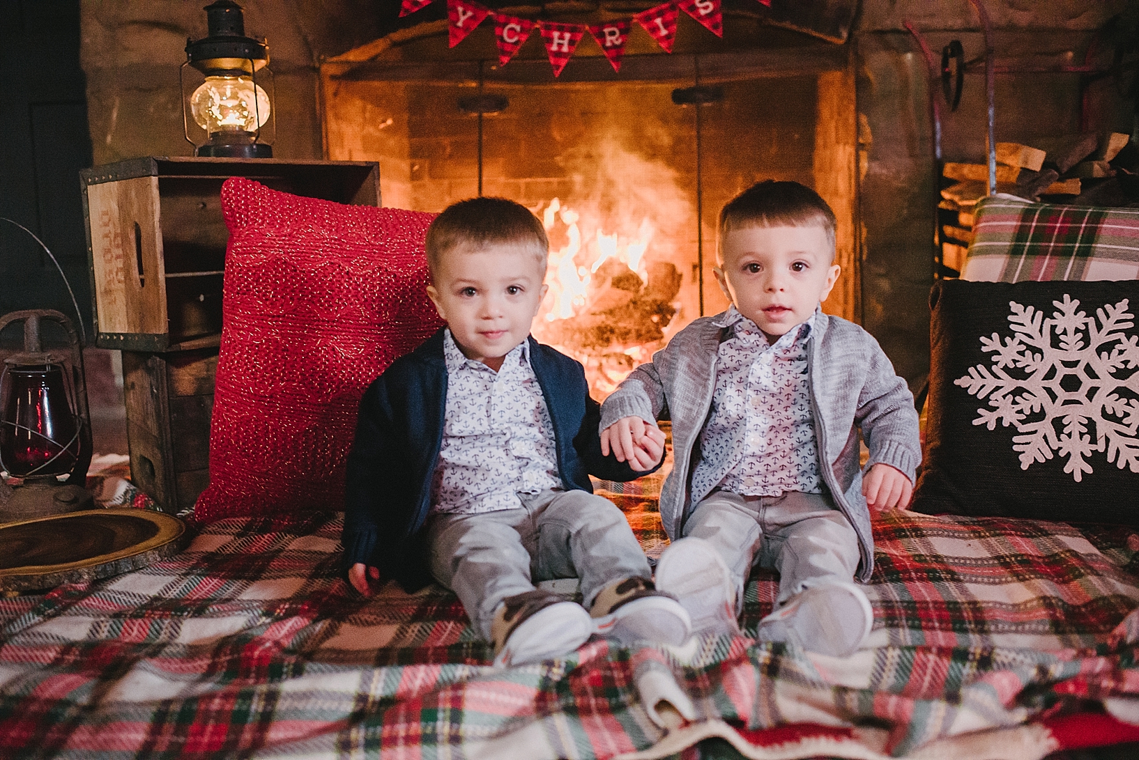 twin toddler boys dressed in navy and grey cardigans sitting on plaid Christmas blanket in front of stone fireplace