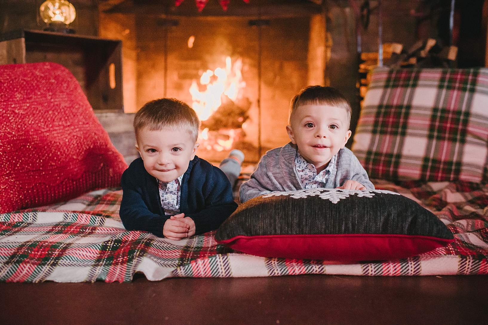 twin toddler boys dressed in navy and grey cardigans laying on plaid Christmas blanket in front of stone fireplace