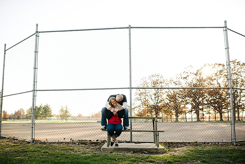 couple sitting on bench in front of baseball field with arms wrapped around each other