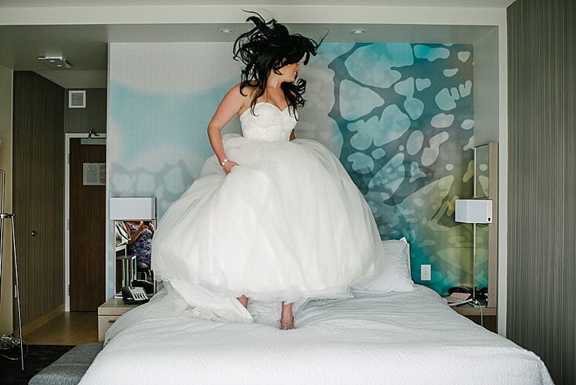 bride jumping on bed in hotel room in her wedding gown