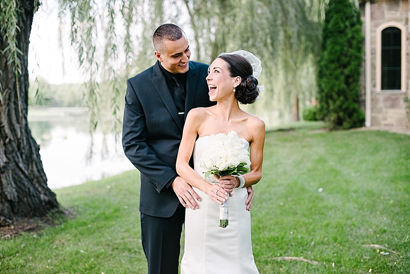bride wearing birdcage veil laughs up at groom in black tux standing in front of willow tree