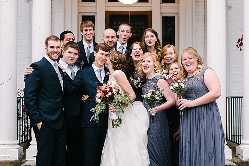 bride kisses groom on cheek as bridal party stands around them laughing and cheering