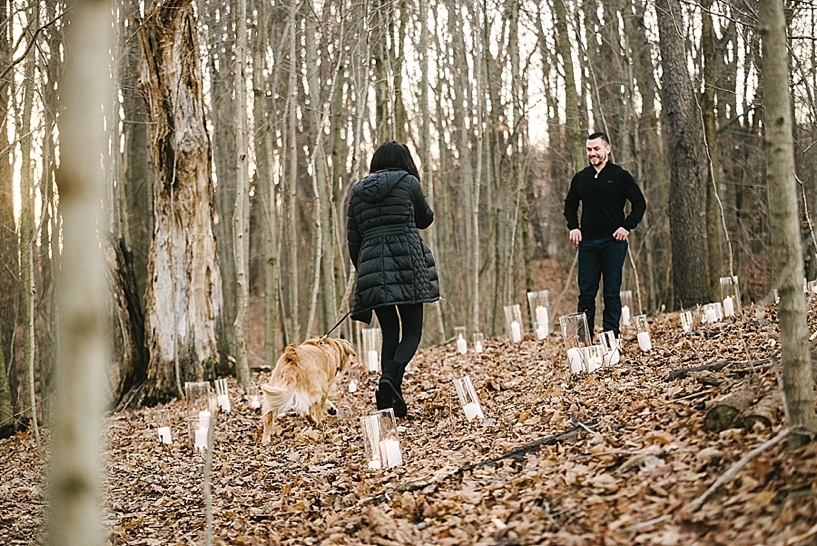 woman in black coat with golden retriever on leash walking up to man in black sweater standing in woods surrounded by candles
