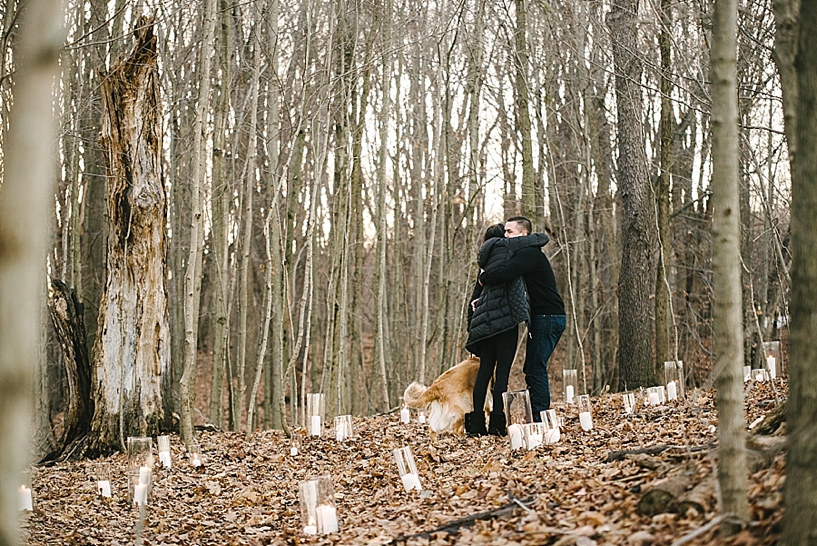 woman in black coat with golden retriever on leash hugging man in black sweater standing in woods surrounded by candles