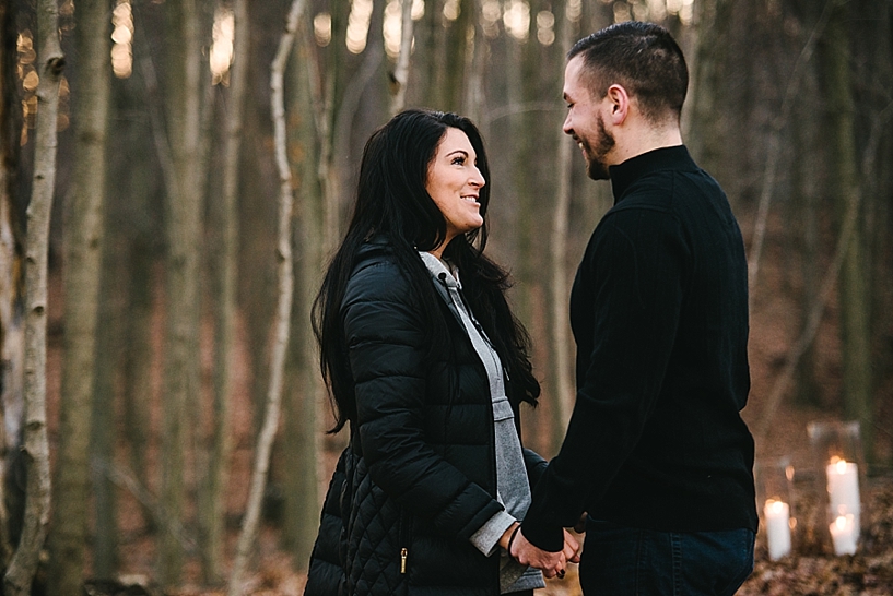 young woman in black coat with black hair smiling at fiance in black sweater in middle of woods