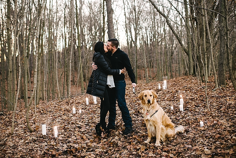 newly engaged couple kissing while holding golden retriever in woods with candles