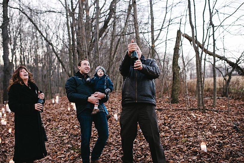 man pops bottle of champagne outside in woods while family looks on